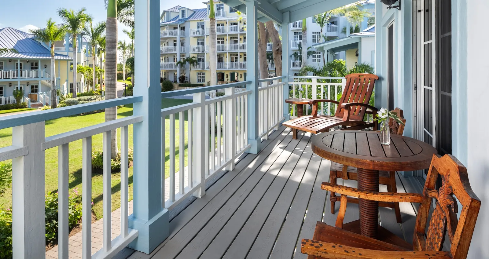 A wooden veranda with teak rocking chairs and a round table, a white railing and the lawn in front.