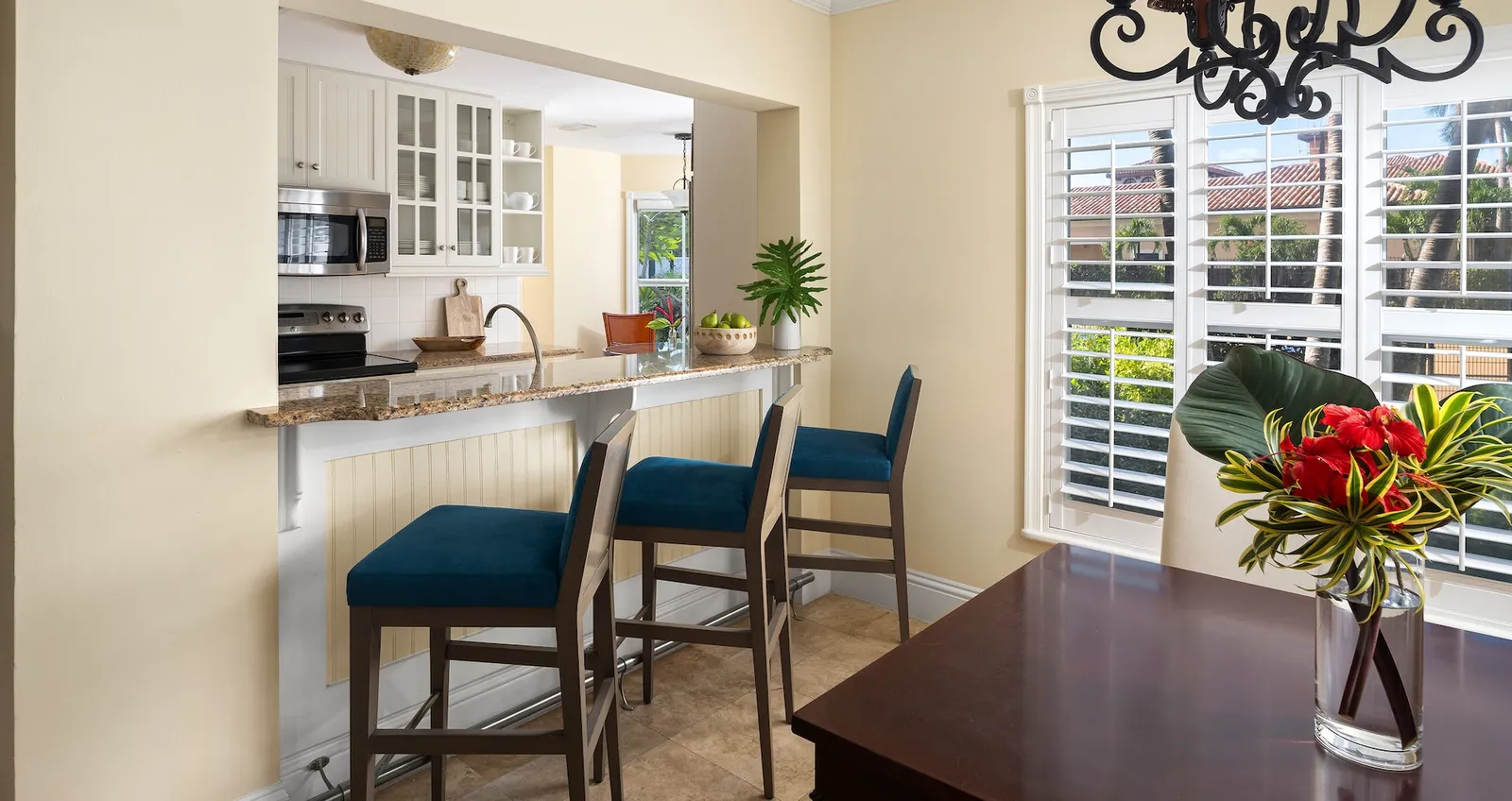 A villa’s kitchen: the granite breakfast bar with blue stools, white shutters and a vase of red flowers on the table.