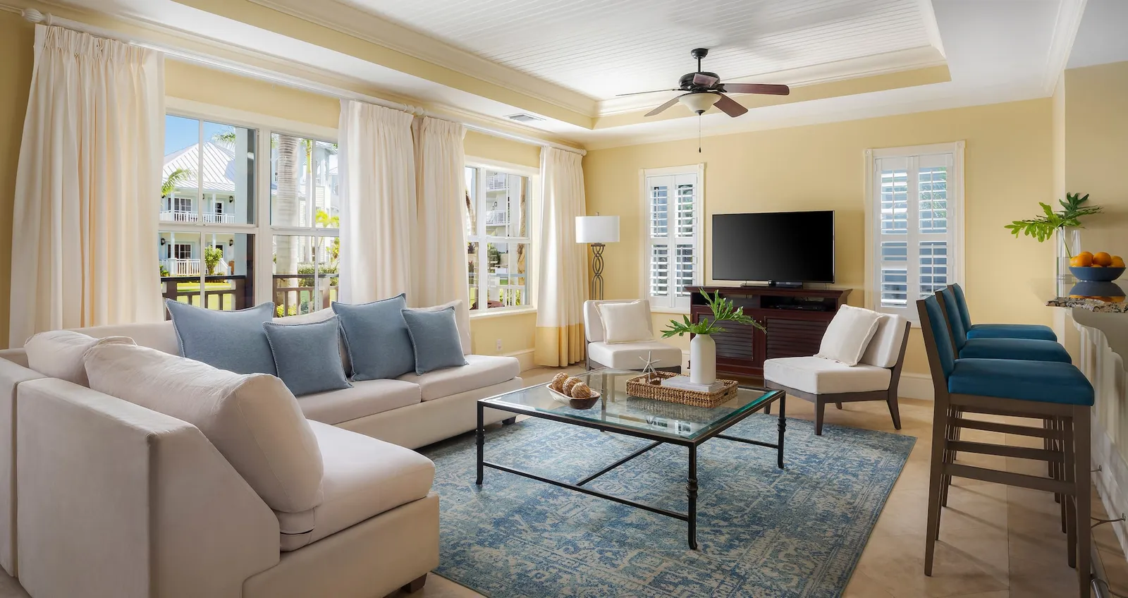 A villa’s living room: a cream corner sofa, the glass coffee table, the television and the blue stools of the breakfast bar.