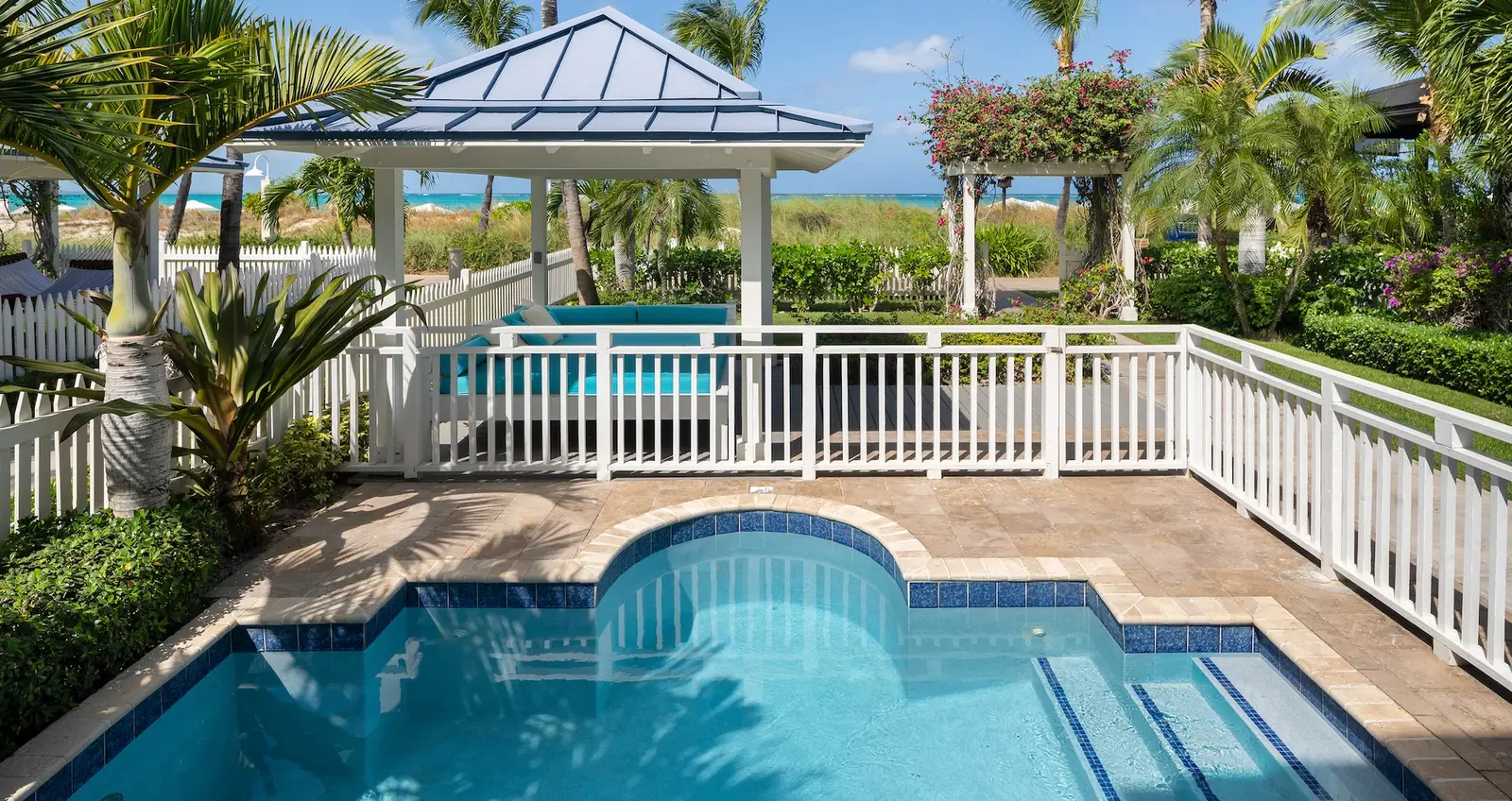 The private pool of a beachfront villa, enclosed by a white fence, with a blue-roofed gazebo and the sea beyond the dune.