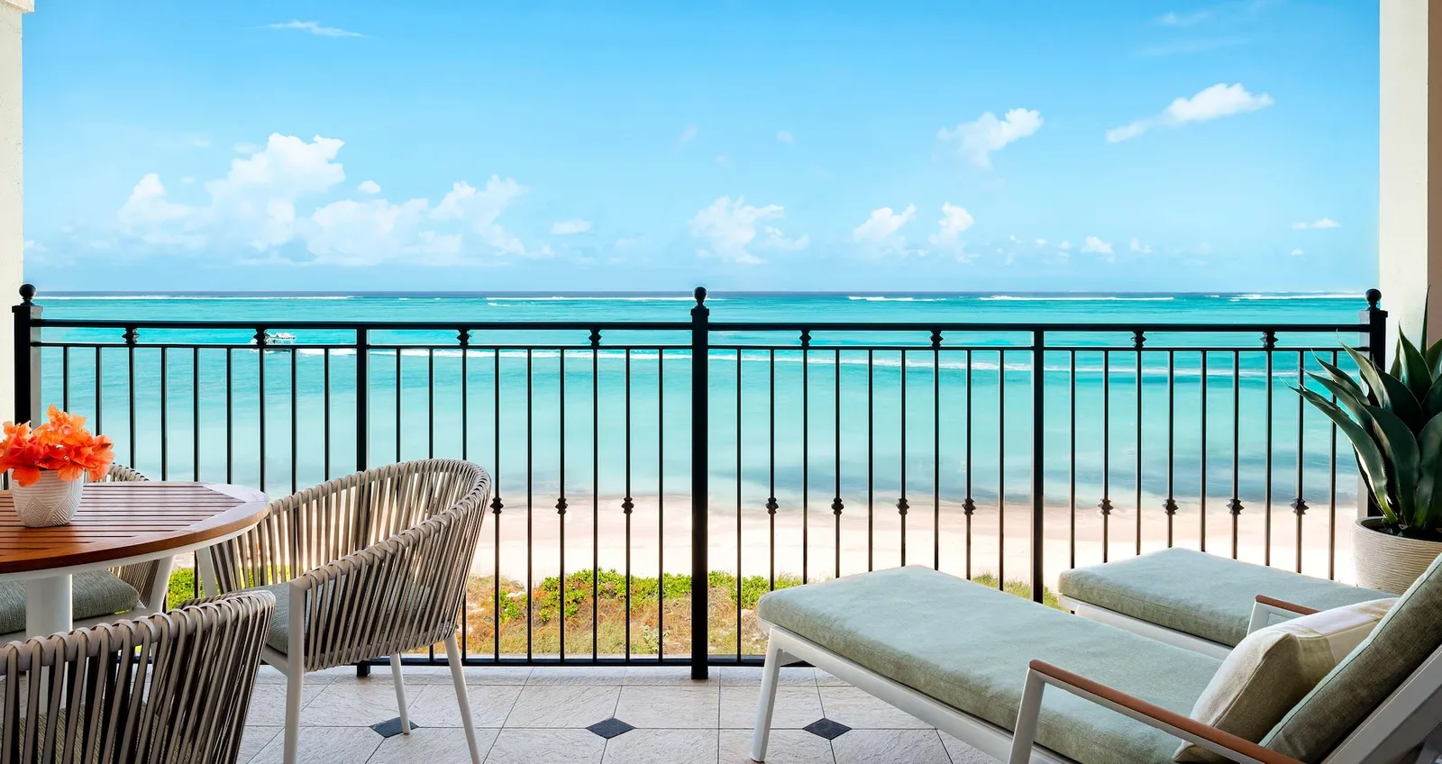 A balcony with a black railing facing the beach and the turquoise sea, with armchairs, a small table and 2 loungers.