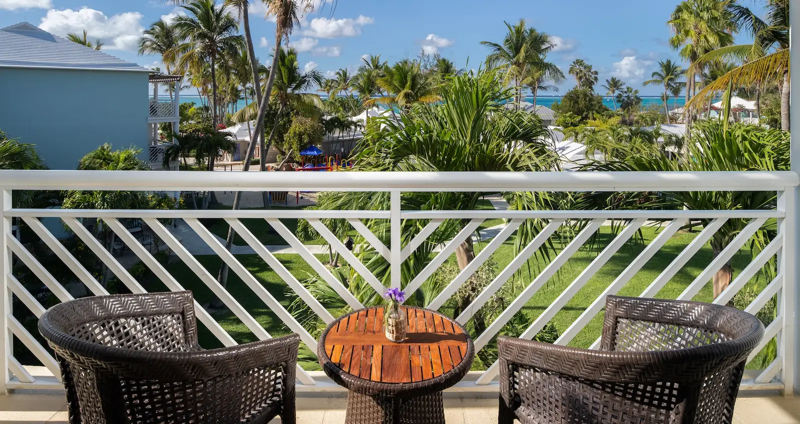 The balcony of a first-floor room: 2 woven armchairs, a small wooden table, a white railing and the garden’s palms.