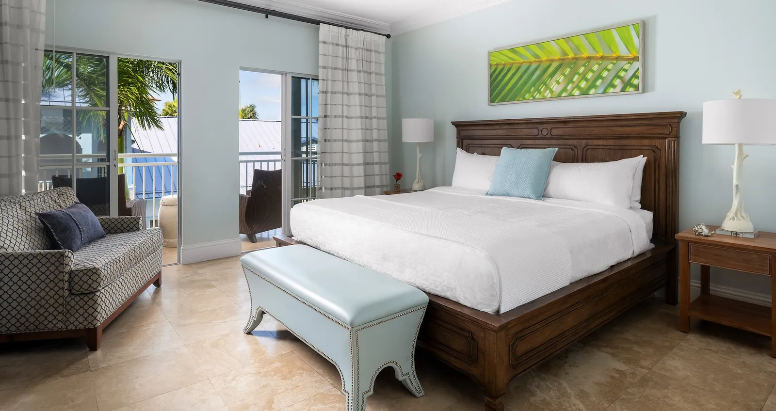 A room with pale blue walls, a king bed, a light blue bench at its foot, an armchair and the doors to the balcony.