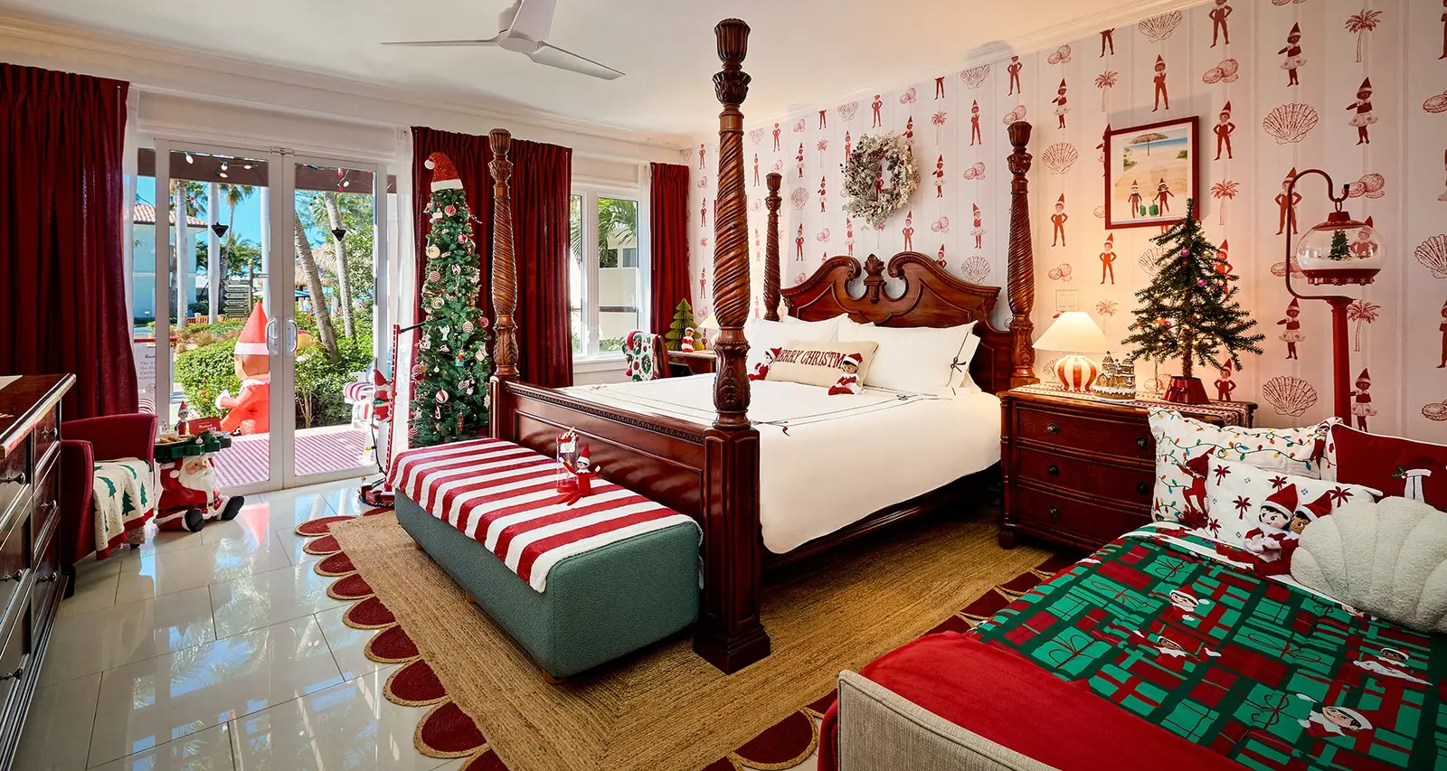 A bedroom dressed for Christmas: the four-poster bed, red-and-white wallpaper with elves, a striped bench and small trees.