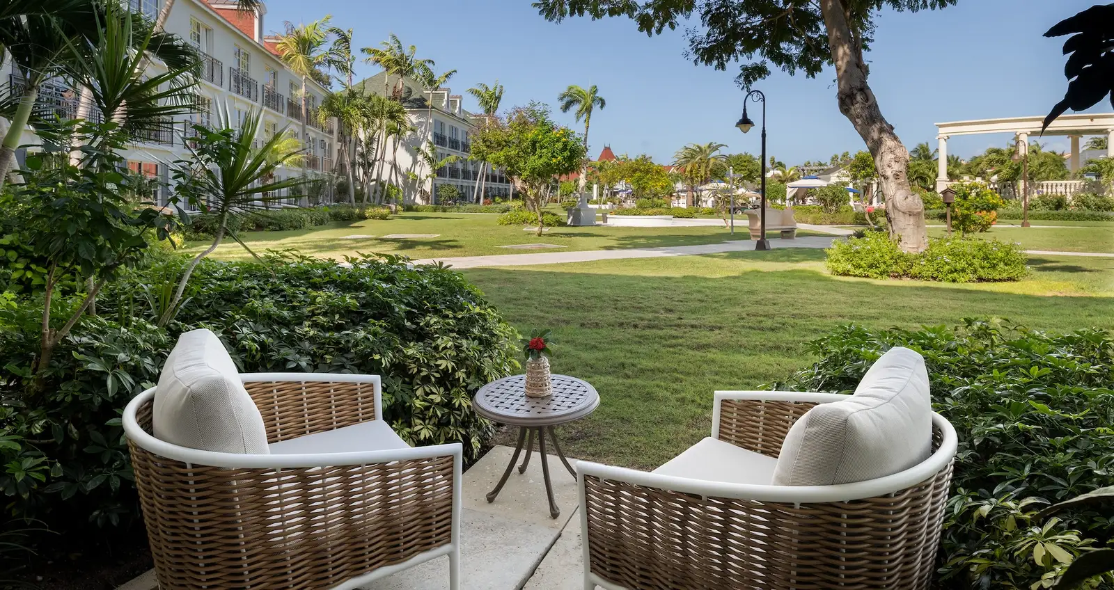 2 rattan armchairs with white cushions on a patio looking over the lawn, palms and the village’s buildings.