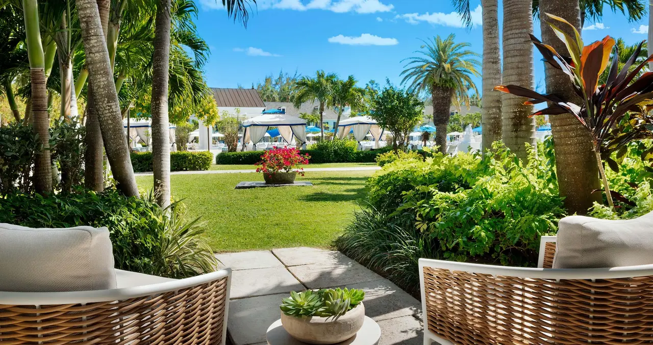 The patio of a ground-floor suite: woven armchairs facing the lawn, the palms and the pool’s cabanas.