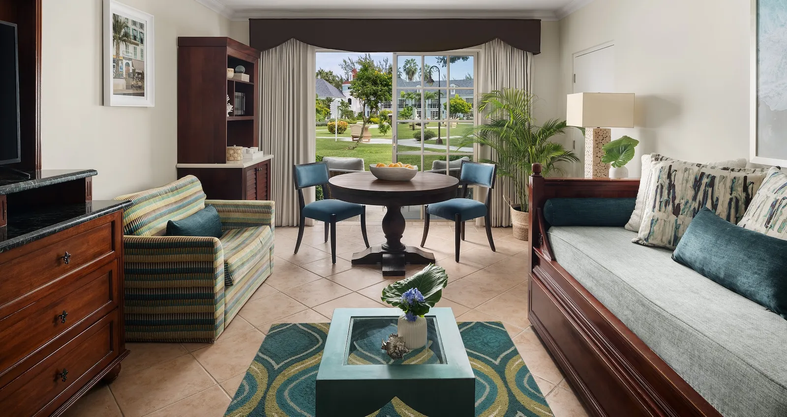A suite’s sitting room: a striped sofa, a dark wooden daybed, the round table with blue chairs and the window onto the lawn.