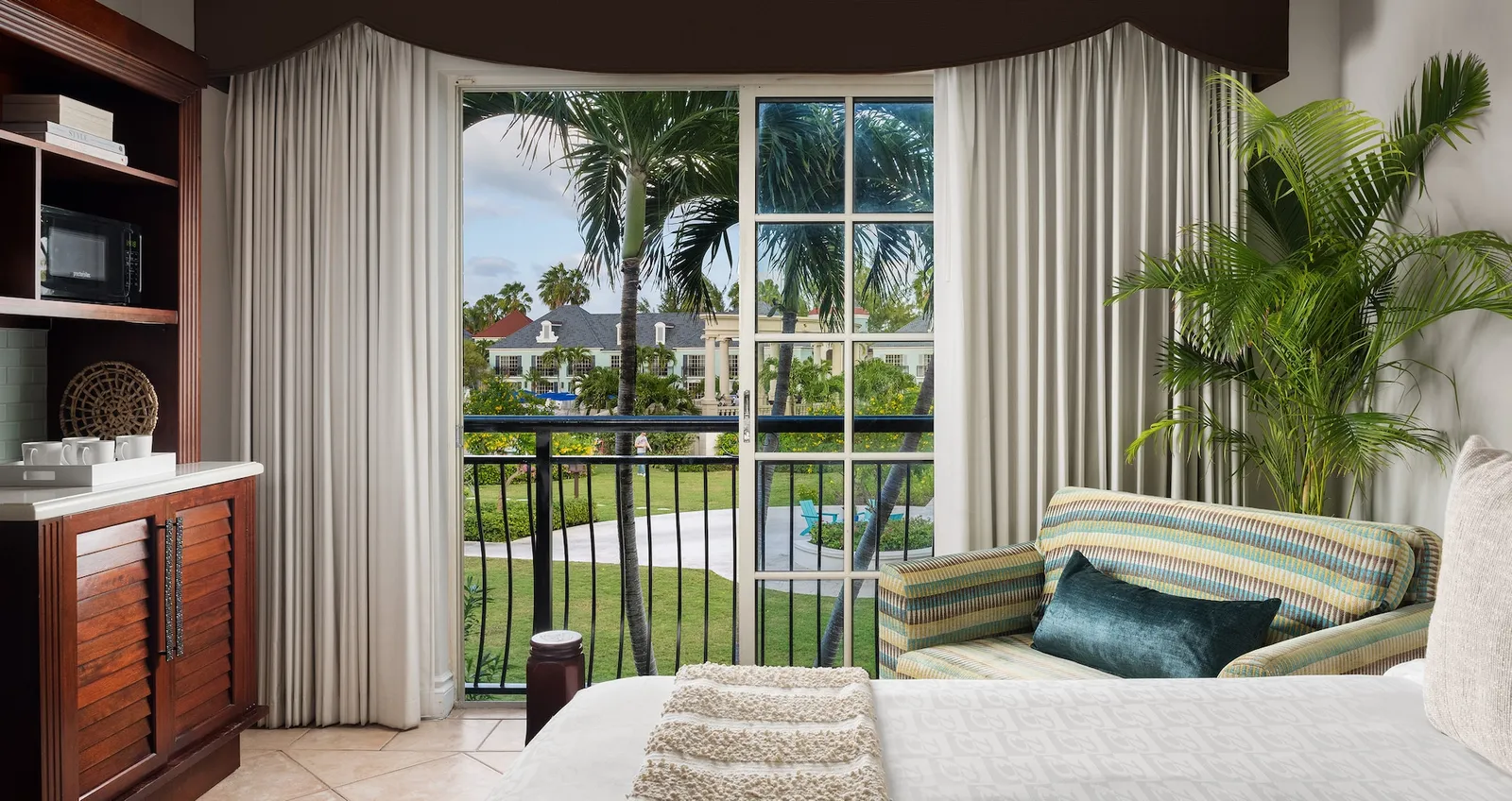From the bed, the French window with its railing open onto palms and the village’s buildings, a striped sofa and a potted palm.
