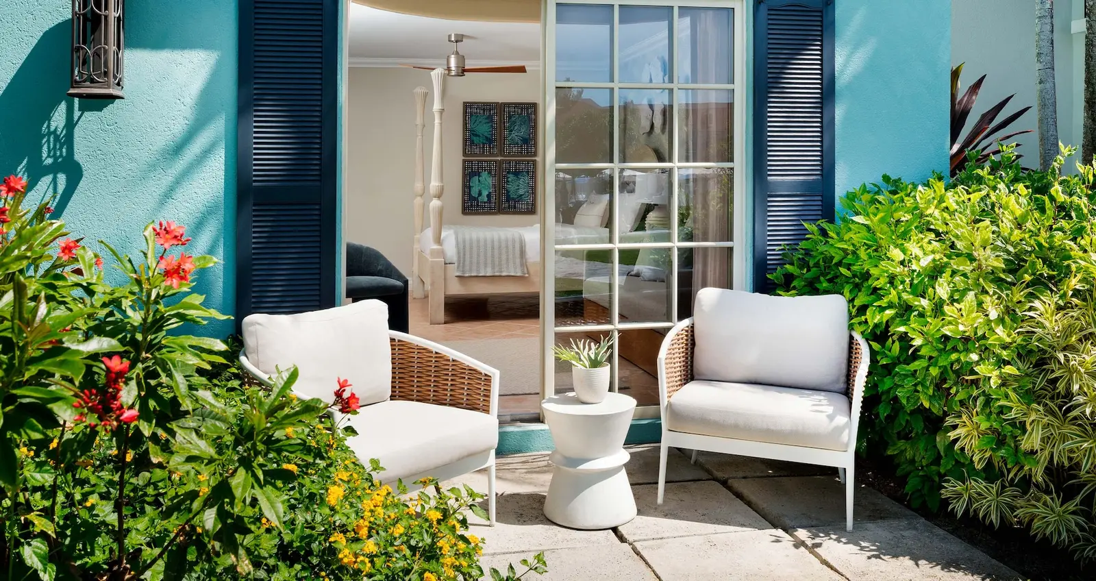 A ground-floor patio: 2 white woven armchairs between flowering shrubs, blue shutters and the bedroom beyond the open door.