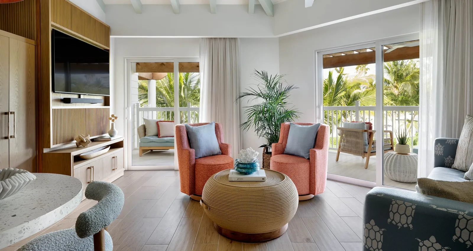 A suite’s living room: 2 coral armchairs, a round woven coffee table, a wooden wall with the television and the doors to the balcony.