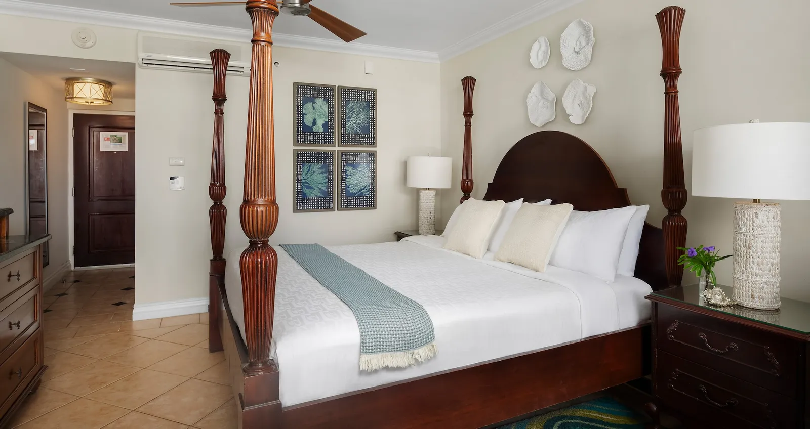 A mahogany four-poster bed with white linen, decorative shells on the wall and a ceiling fan.