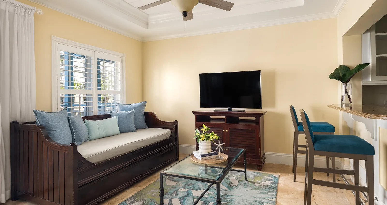 A living room with a dark wooden daybed and blue cushions, the television, a glass coffee table and a breakfast bar with 2 stools.