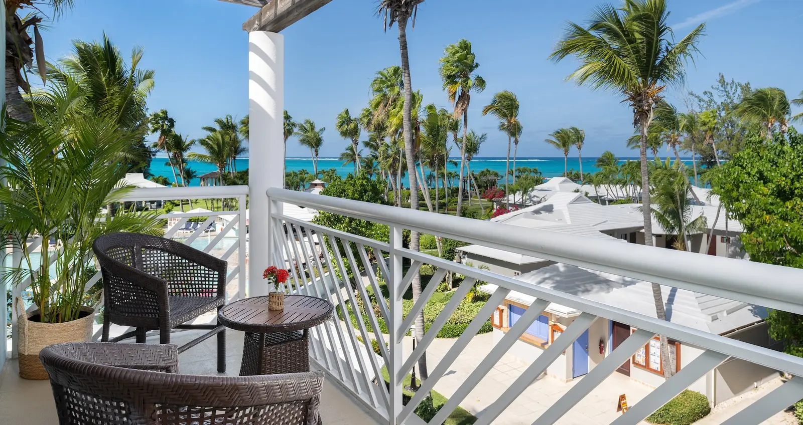 A balcony with woven armchairs and a white railing, above roofs and palms, with the turquoise sea in the distance.
