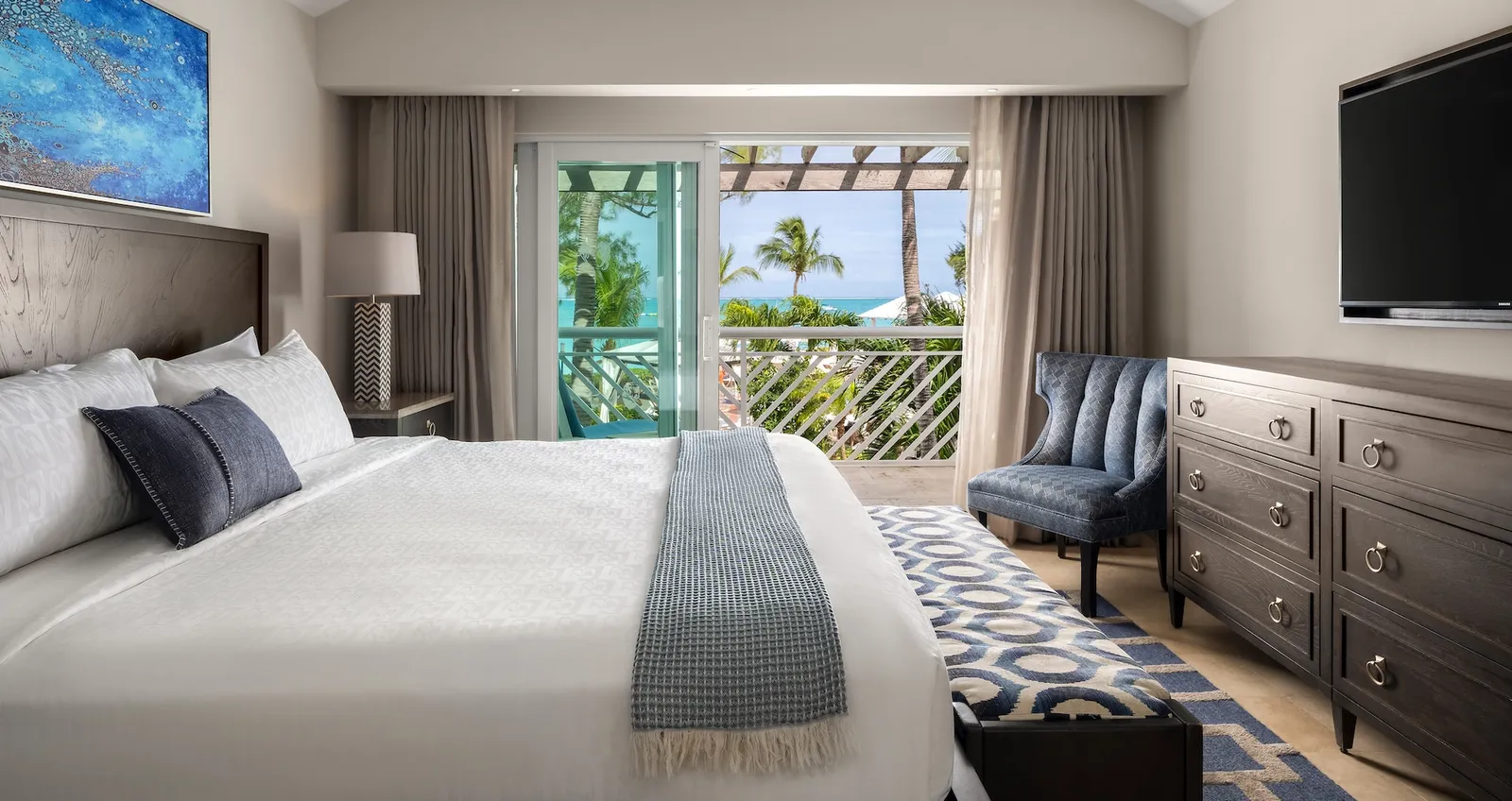 A pale bedroom with a king bed, a grey chest of drawers with the television, an armchair and the door to a patio among palms.
