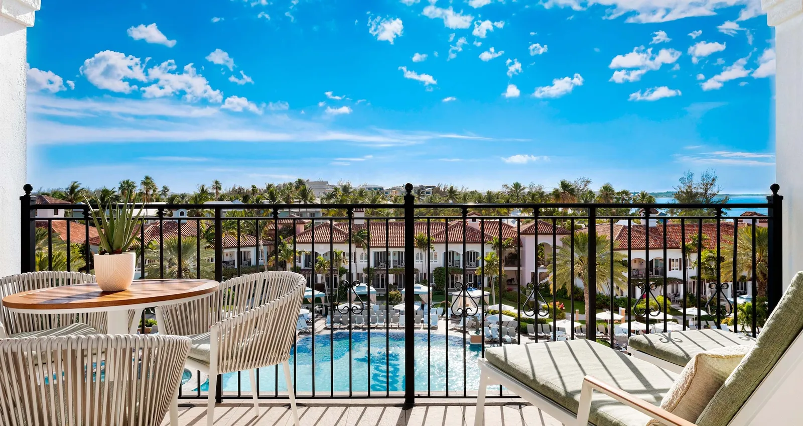 A top-floor balcony with a table, white chairs and loungers, above the pool and the village’s red roofs.
