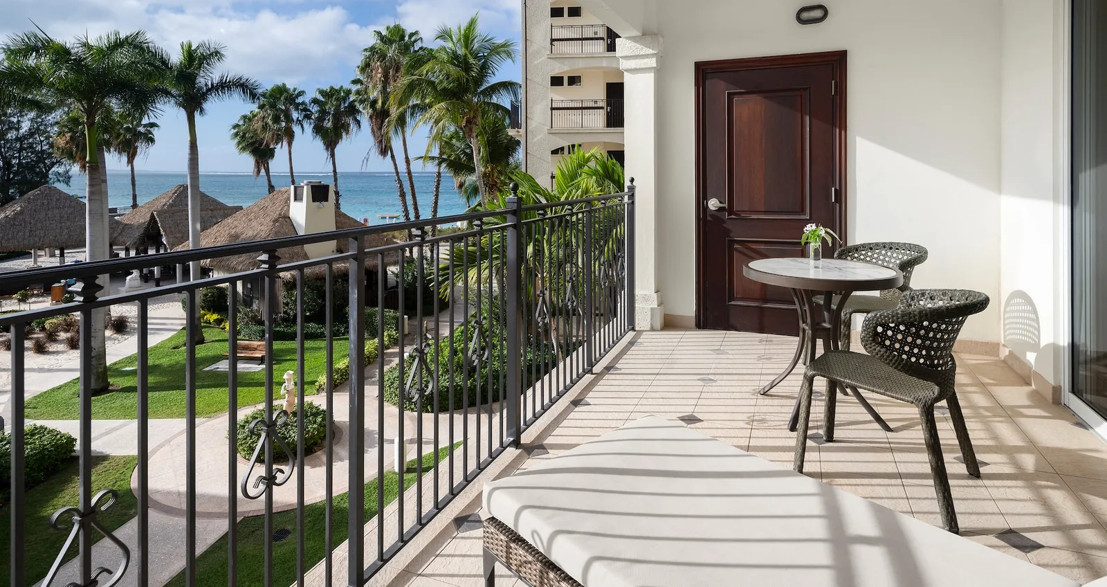 A long balcony with a black railing, a small table with 2 chairs and a lounger, above paths and palms, with a glimpse of the sea.