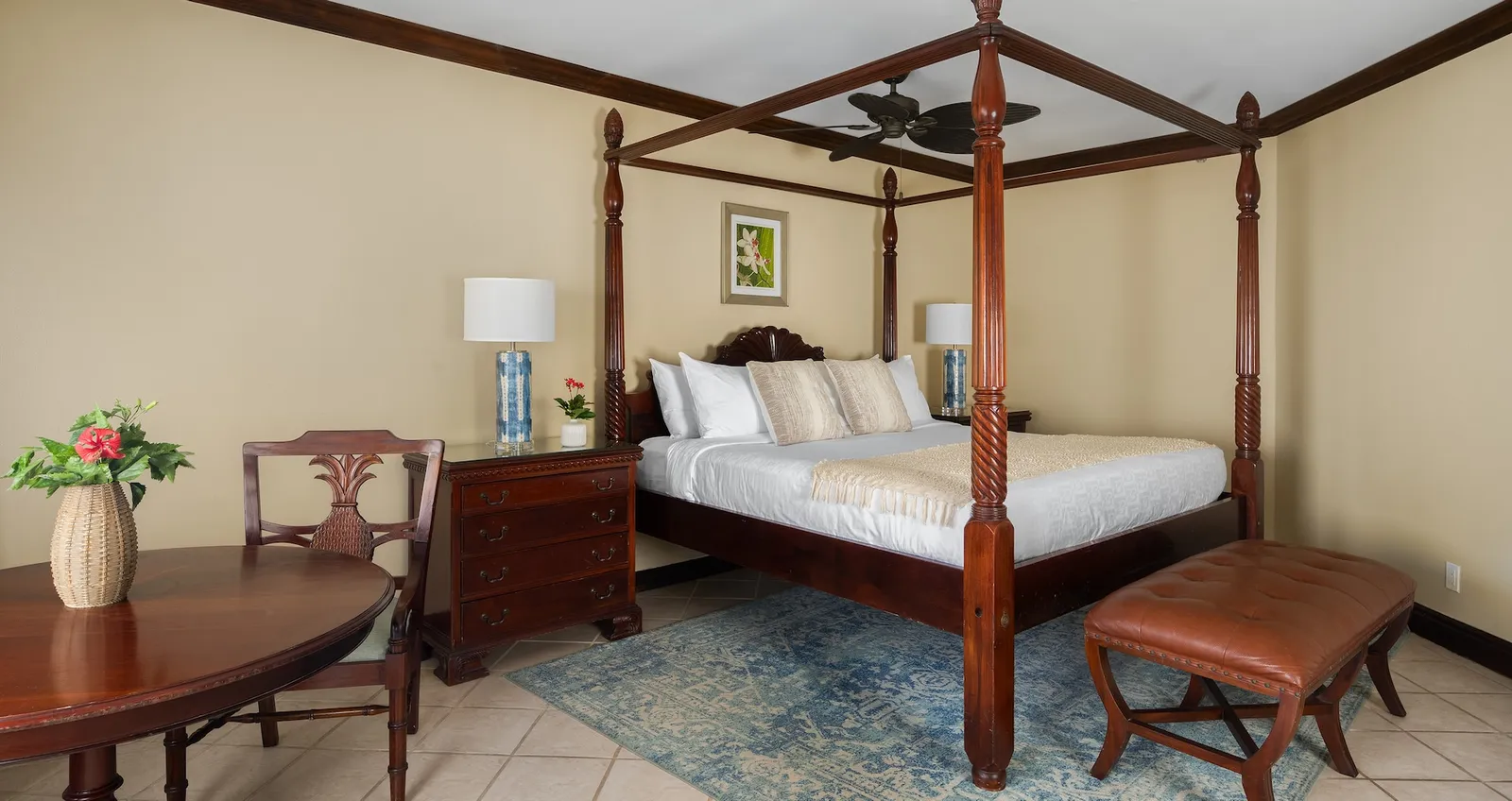 A mahogany four-poster king bed, a round table with flowers, a leather bench and a pale tiled floor.