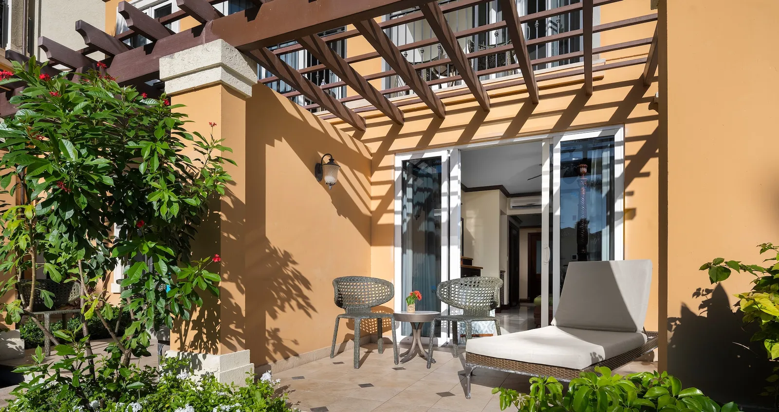 A ground-floor terrace under a wooden pergola, between ochre walls, with a lounger, 2 chairs and a shrub in flower.