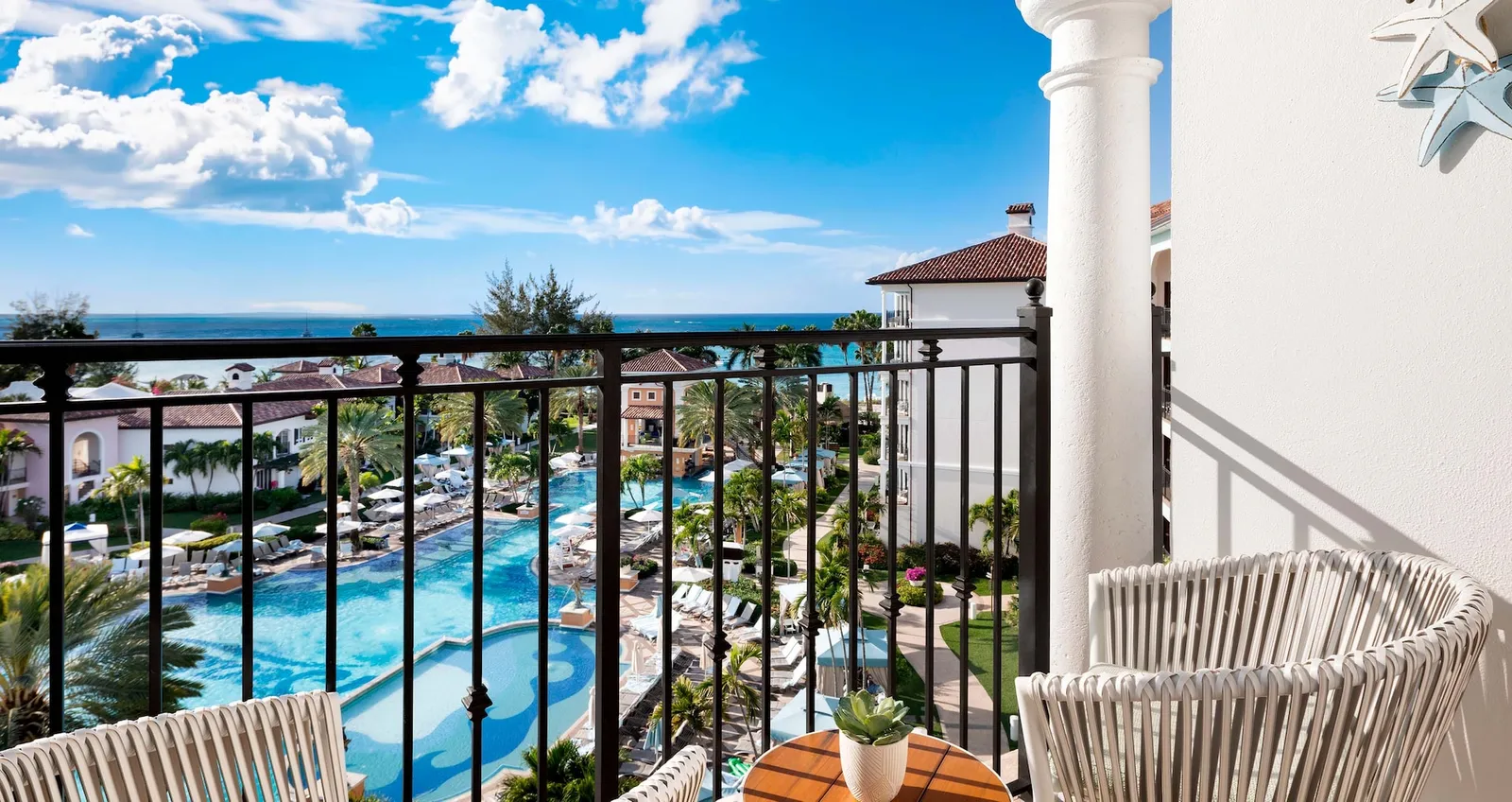 A top-floor balcony above the Italian Village pool, with white armchairs and the sea on the horizon.