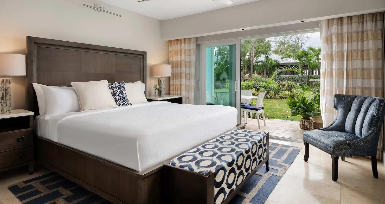 A king bed with a tall wooden headboard, a blue-patterned bench, an armchair and the sliding door to a patio and lawn.