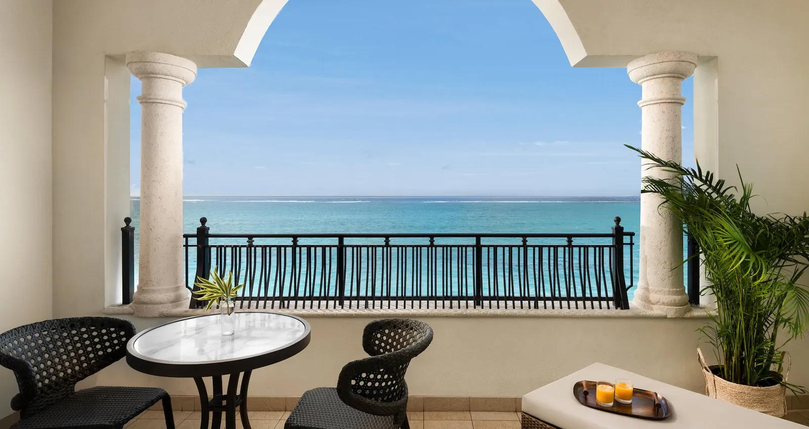 A terrace whose 2 columns and arch frame the sea, with a round table, dark chairs and a potted palm.