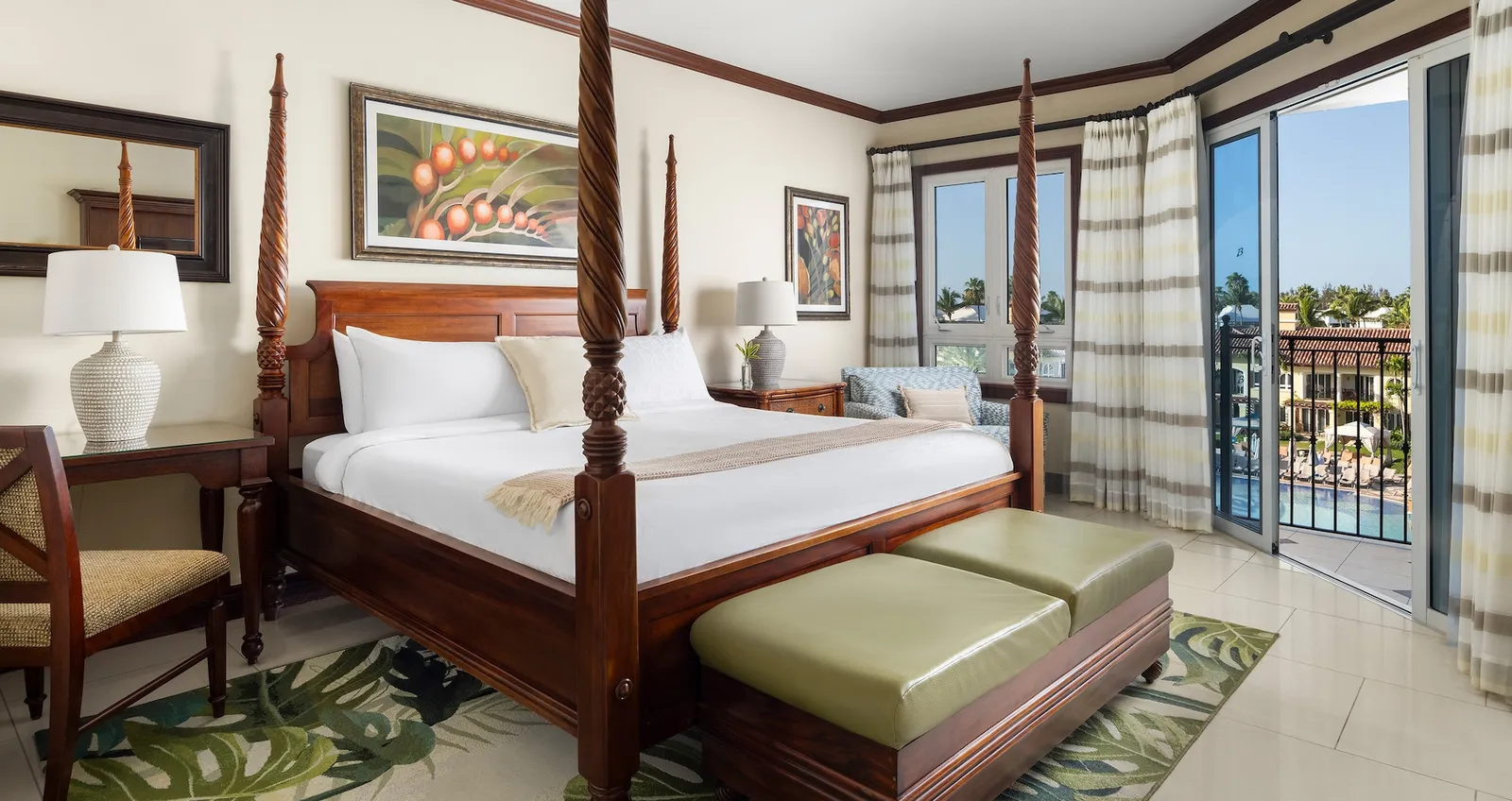 A four-poster king bed with 2 green ottomans at its foot and, beyond the door, the balcony above the pool.