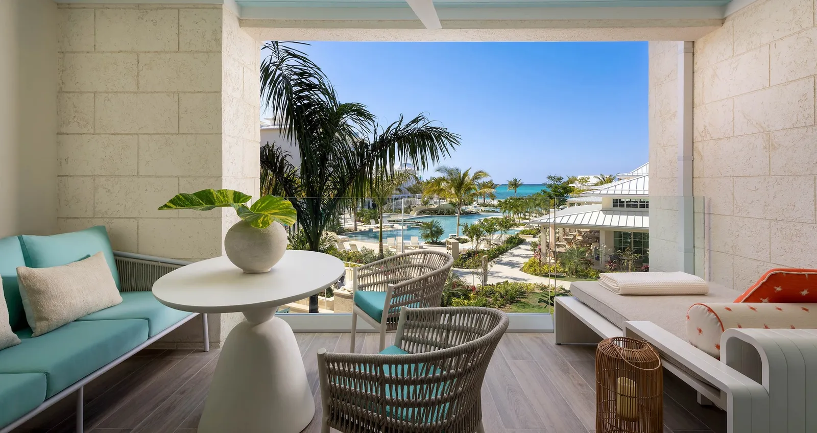 A fourth-floor balcony with a turquoise sofa and a small table, above the lagoon pool, the palms and the sea.