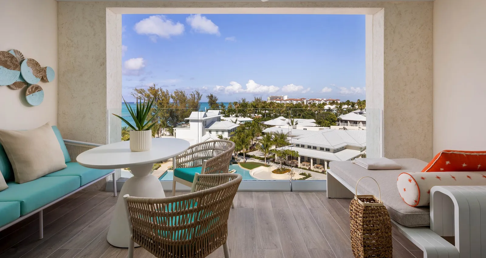 A balcony with a sofa, a round table and a woven armchair, with a view over white roofs to the sea.