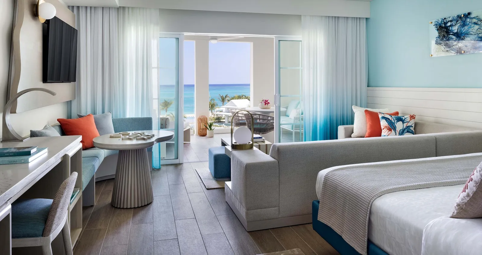 A bedroom opening onto a sitting area and a balcony facing the sea, with blue ombré curtains and coral cushions.