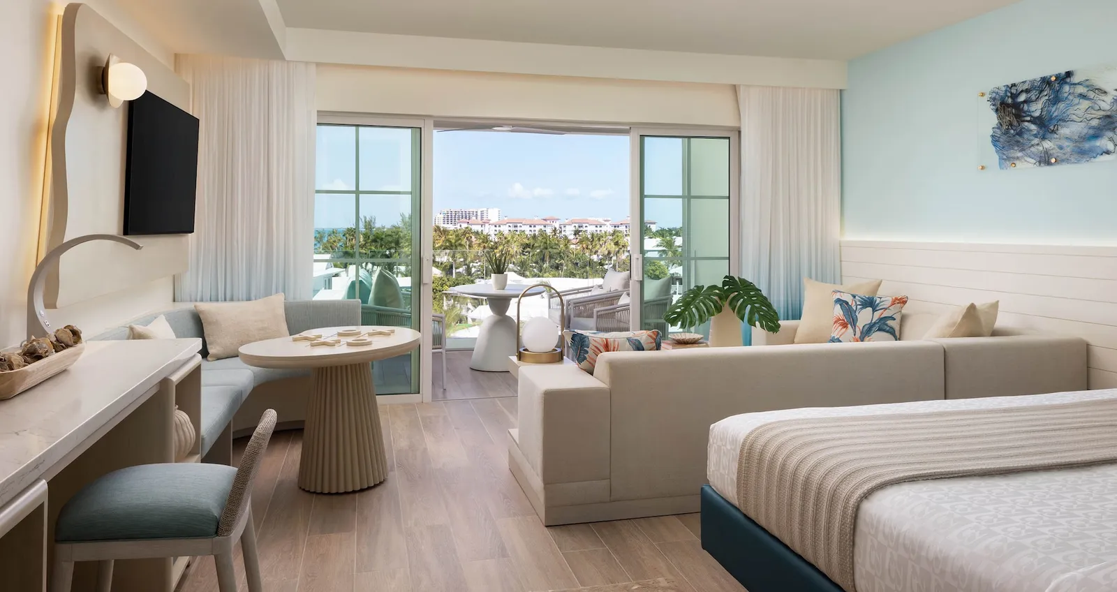 A bedroom with a sitting area: a pale sofa, a round table, a sky-blue wall and the door to a balcony above the palms.
