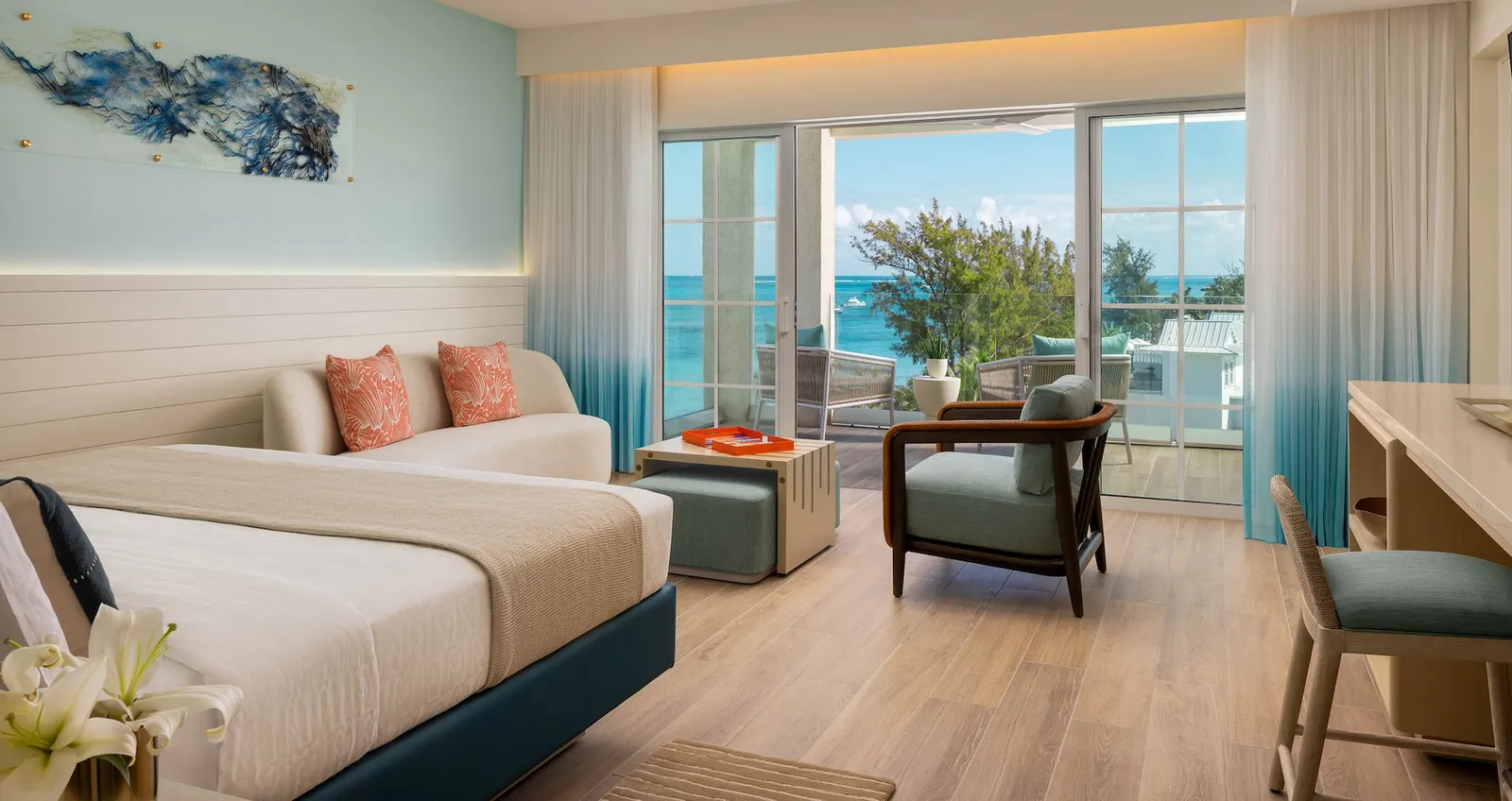 A bedroom with a sofa at the foot of the bed, an armchair, blue ombré curtains and glass doors to the balcony and the sea.