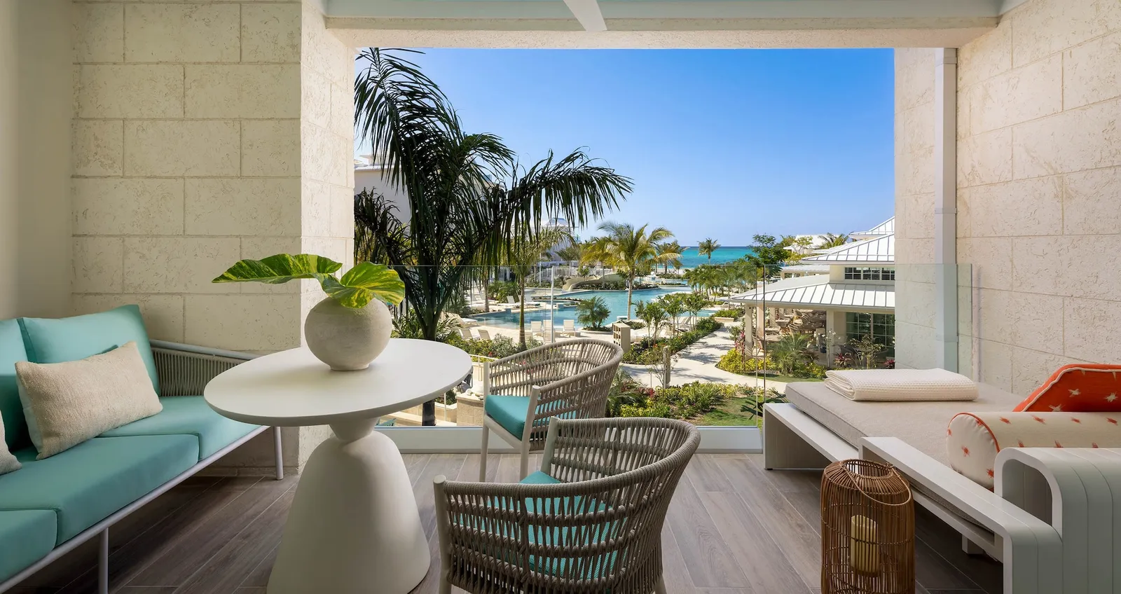 A balcony between 2 pale stone walls, with a sofa and a small table, looking over palms and the lagoon pool.