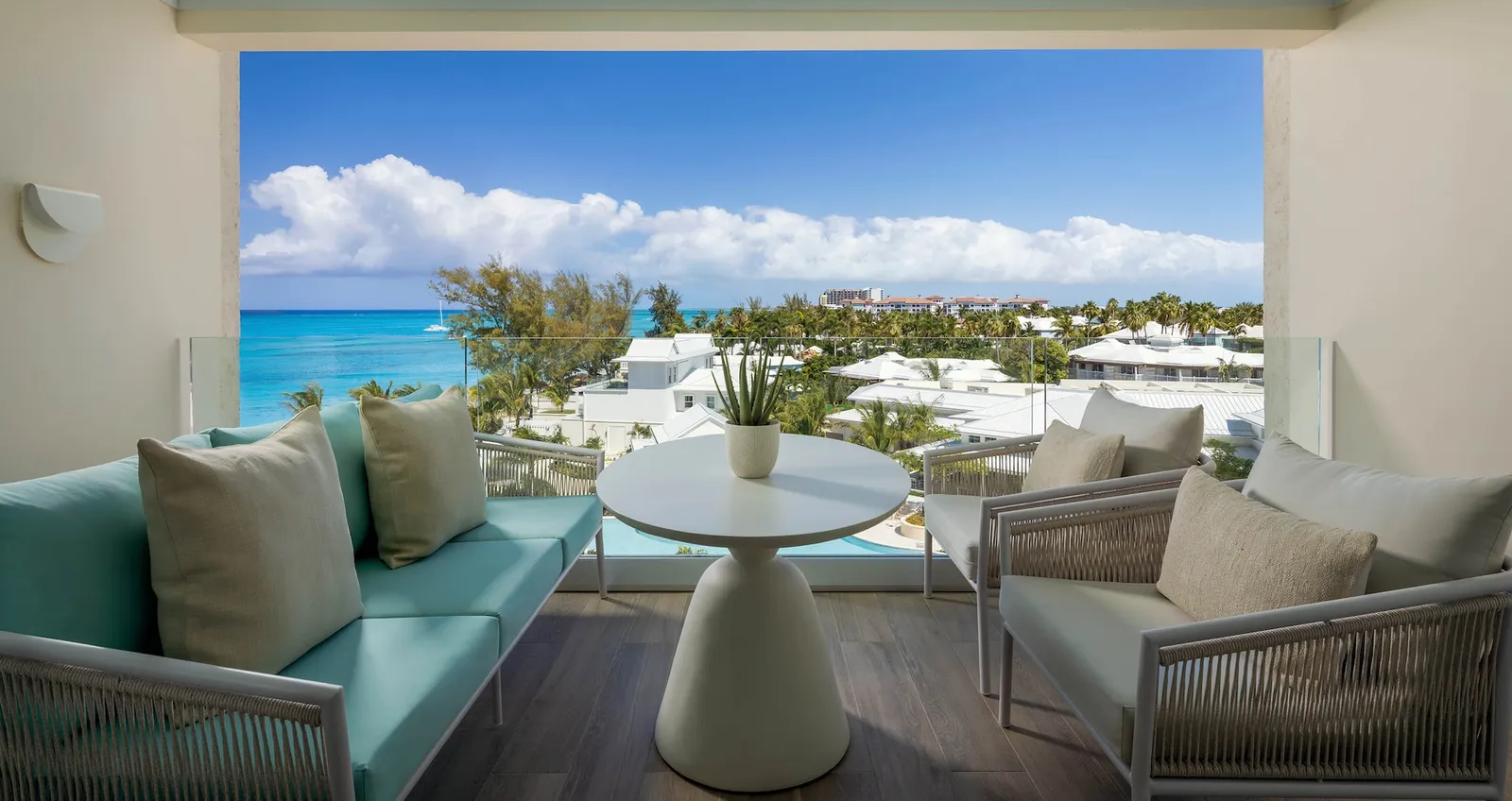 A balcony with a turquoise sofa, a round white table and an armchair, above white roofs, palms and the sea.