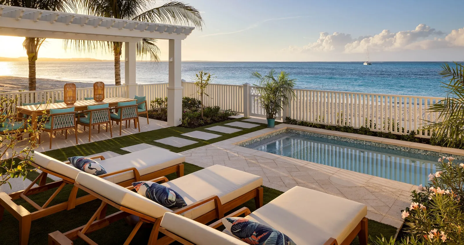A villa’s terrace at sunset: the private pool, loungers, a lawn with stepping stones, the table under a pergola and the beach beyond a white fence.