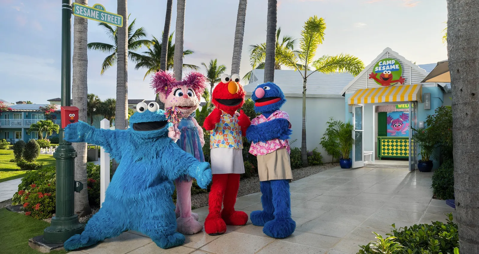 4 Sesame Street characters in costume wave from a path, in front of a brightly painted kiosk and palms.