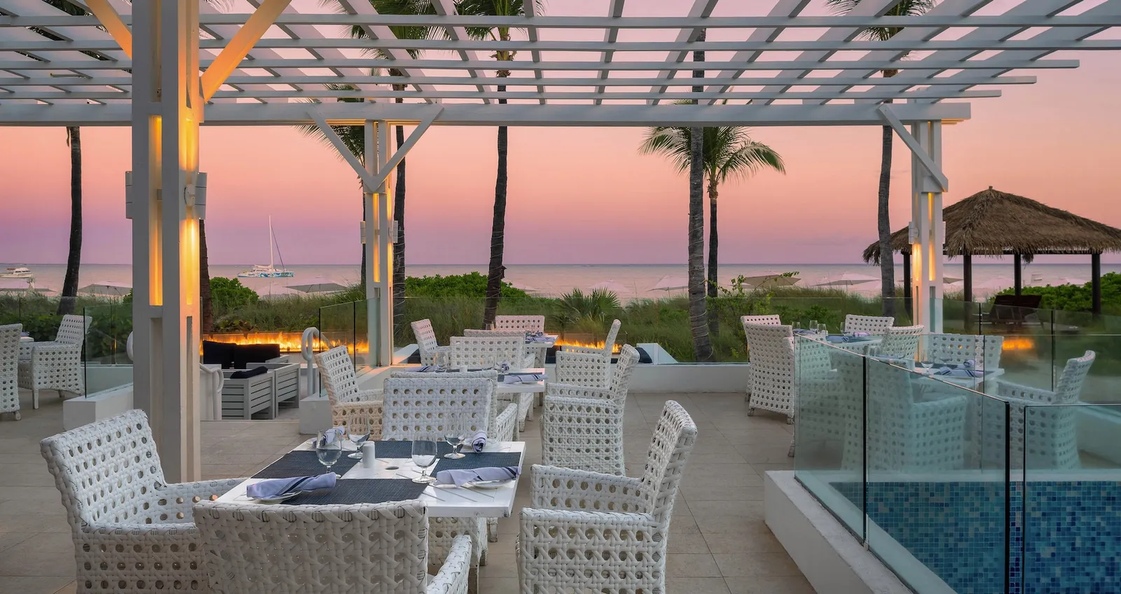 A terrace at sunset: white woven chairs and tables under a white pergola, with palms and a pink sea behind.