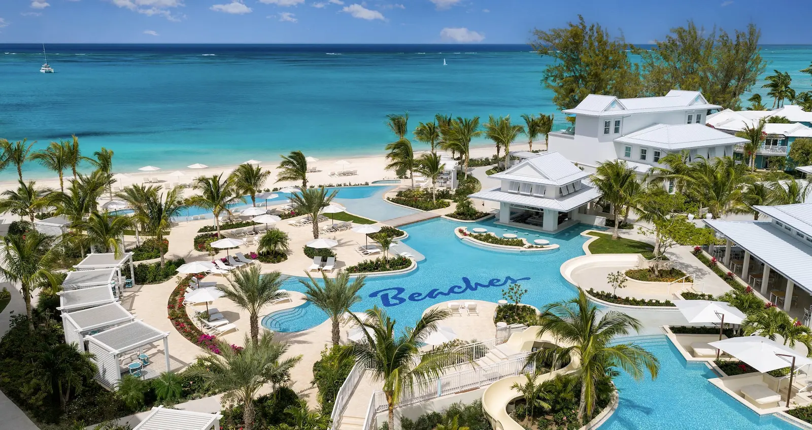 From above, the lagoon pool of Treasure Beach Village with the word Beaches on its floor, among palms, white cabanas and pale-roofed buildings, with the sea behind.