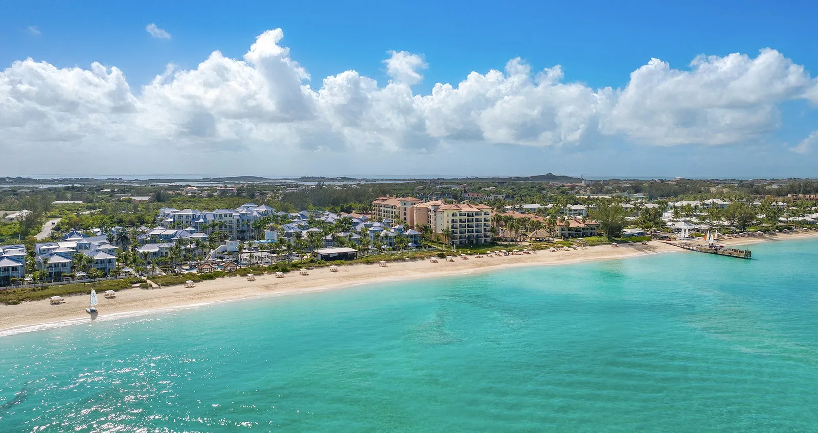 The coast from above the sea: a long white beach, shallow turquoise water, the resort’s roofs among the trees and low clouds over the flat island.
