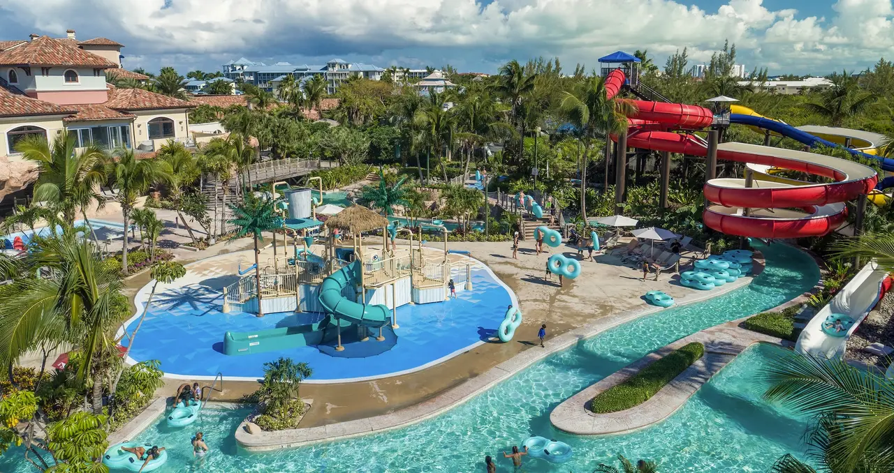 The water park from above: a pool with a play structure at its centre, the lazy river winding around it and the red slides behind.