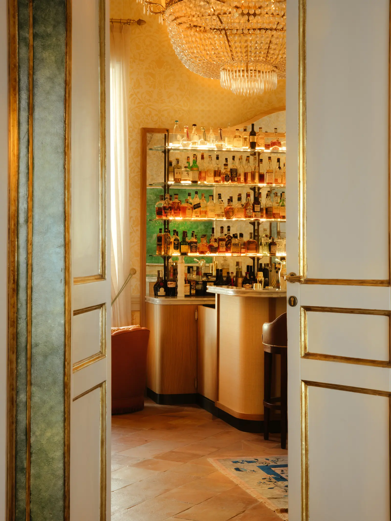 The counter of the Adagio bar seen through a half-open gilded door, with lit bottles and a crystal chandelier.