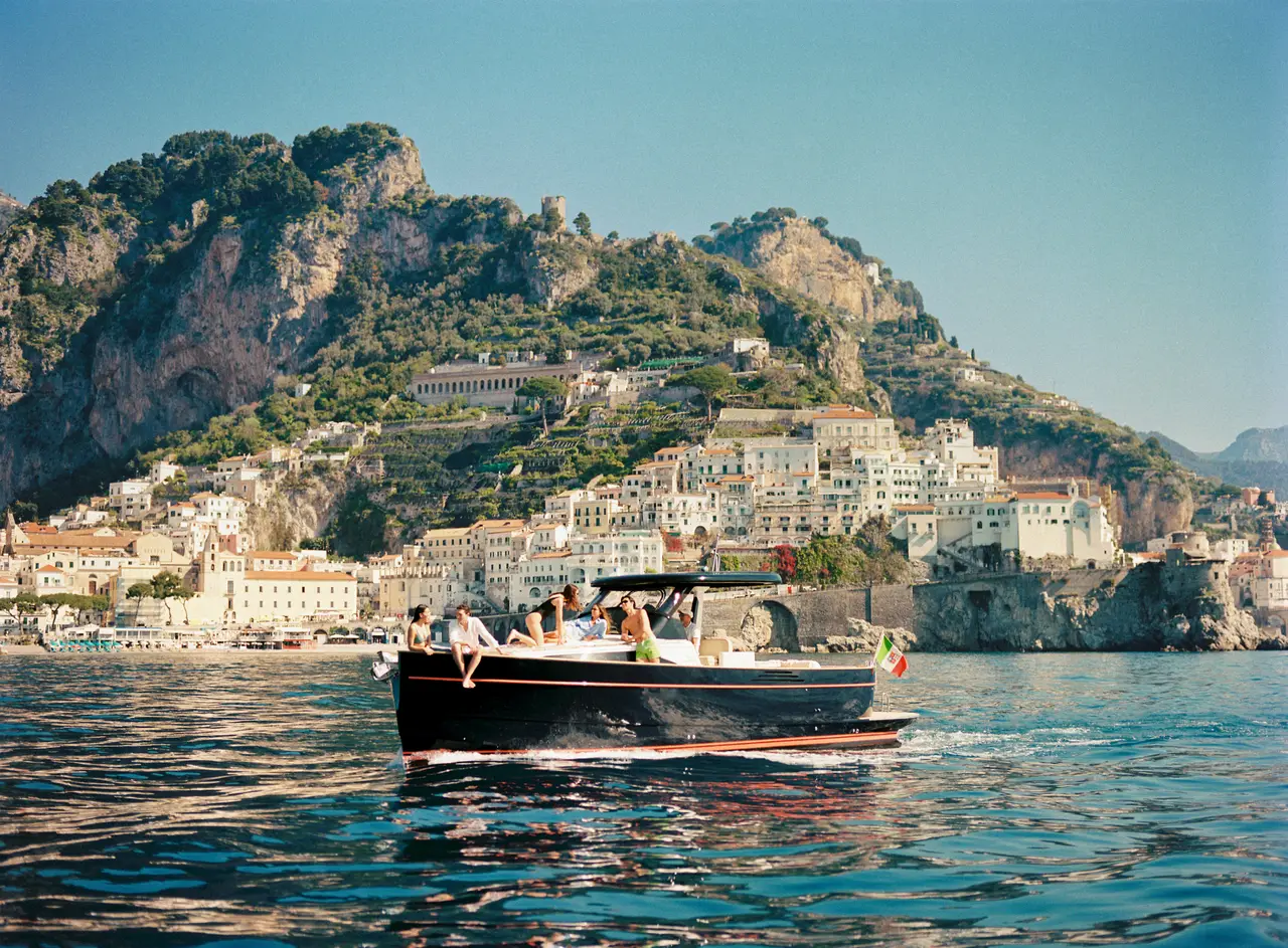 A dark wooden boat with a few guests aboard before the white houses of a coastal town, under the mountain.