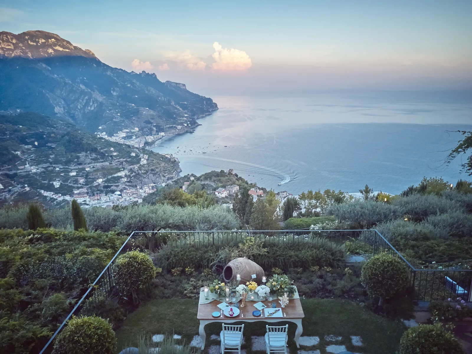 A table for 2 laid in the garden, with the coast and the sea behind at dusk.