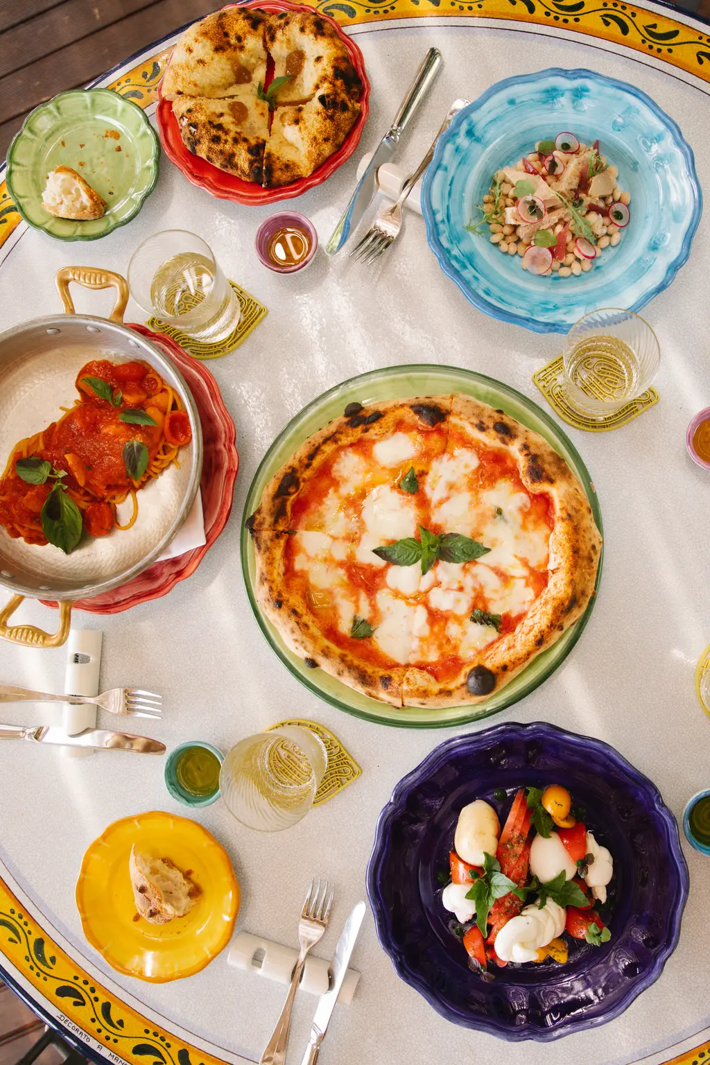 A table seen from above, with a margherita pizza and bright ceramic plates of starters.
