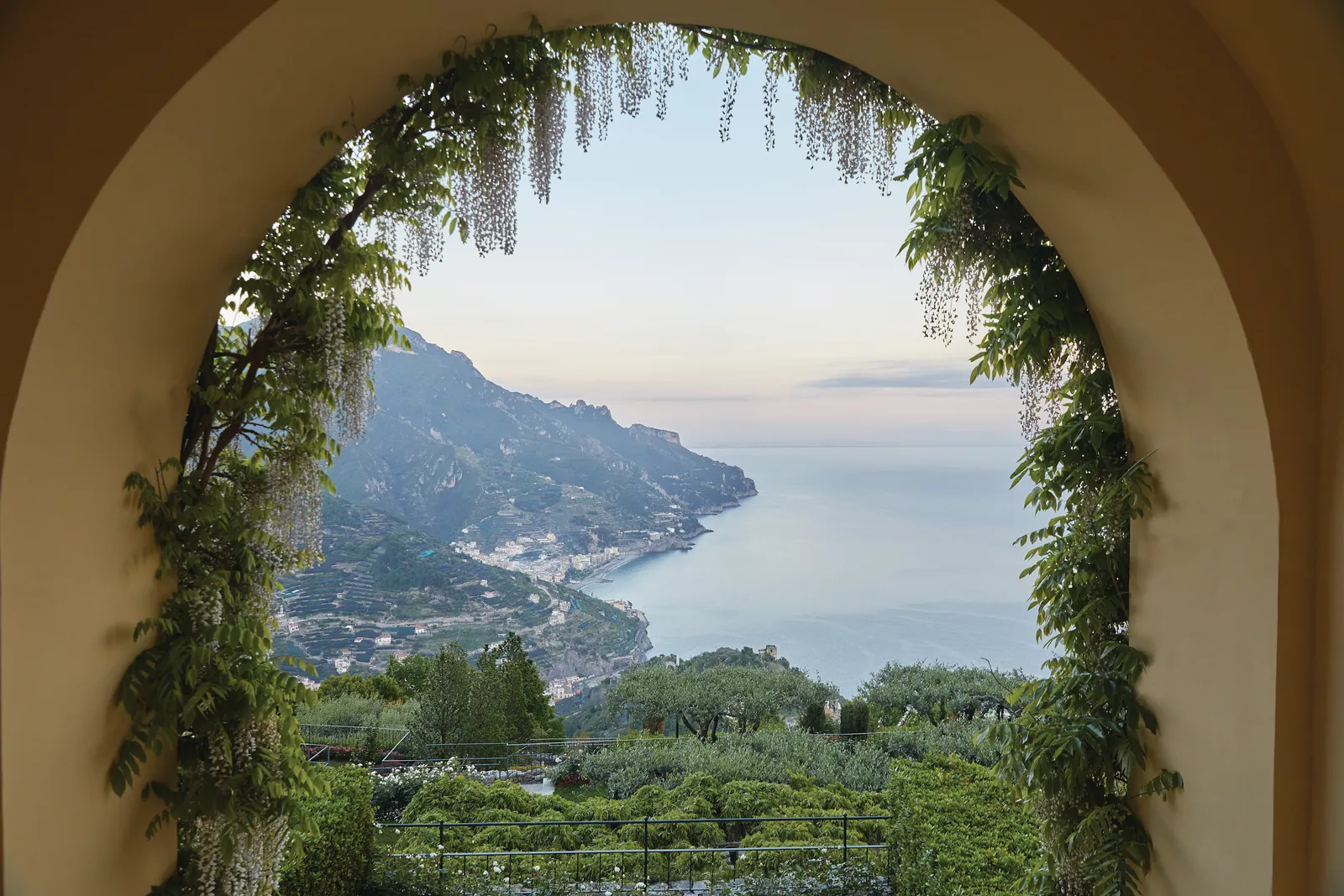 An arch draped in wisteria frames the coast and the sea at sunset, above green terraces.