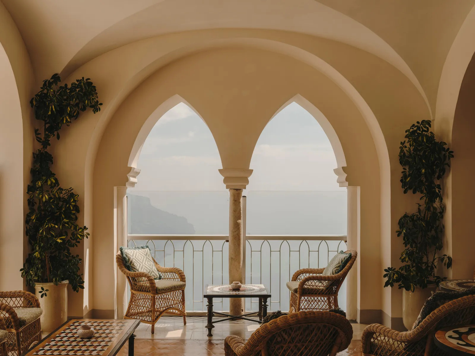 The loggia with its pointed twin arches open to the sea: wicker armchairs, a low table and plants on either side.