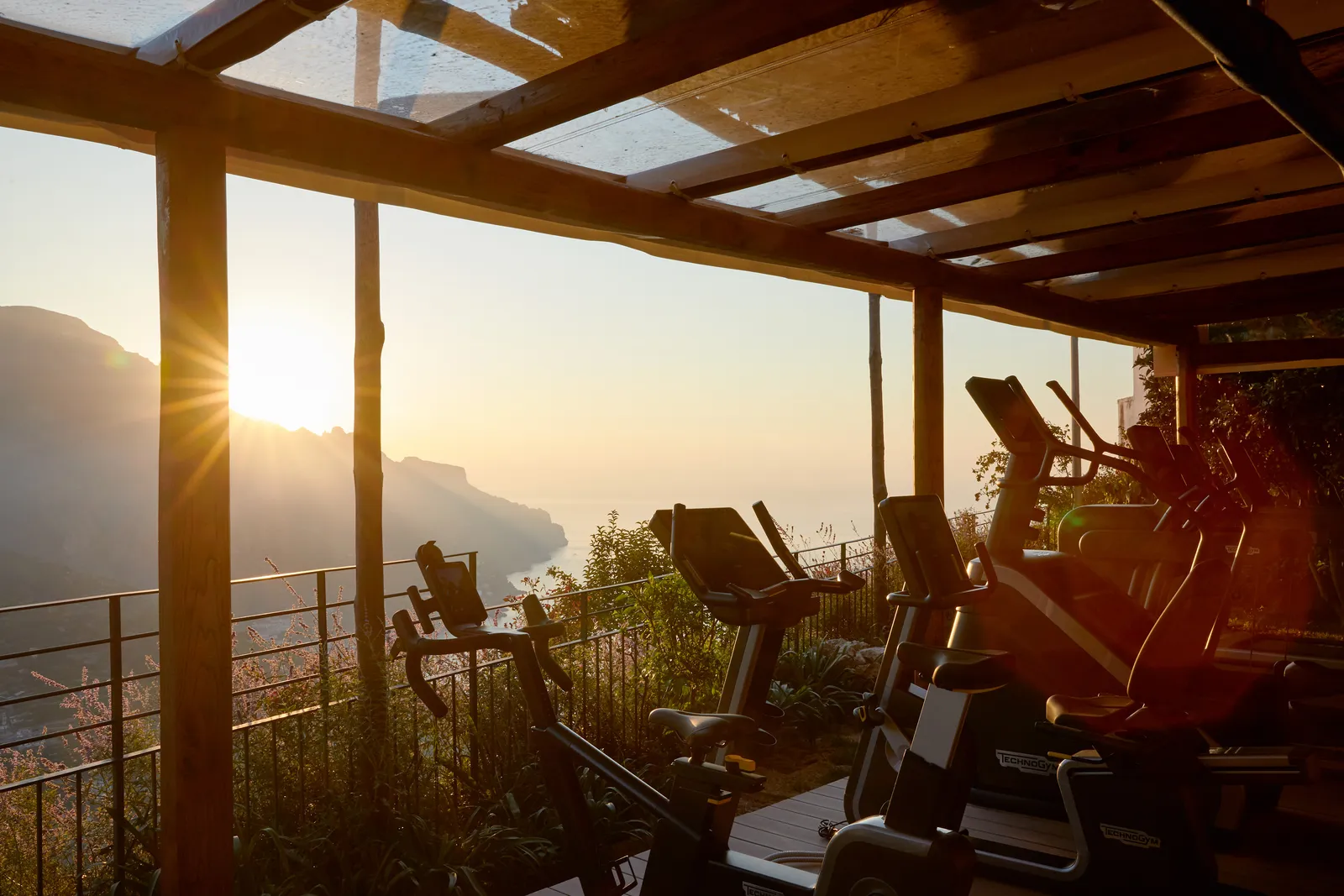 The open-air gym under a wooden pergola: cardio machines facing the low sun over the mountains and the sea.