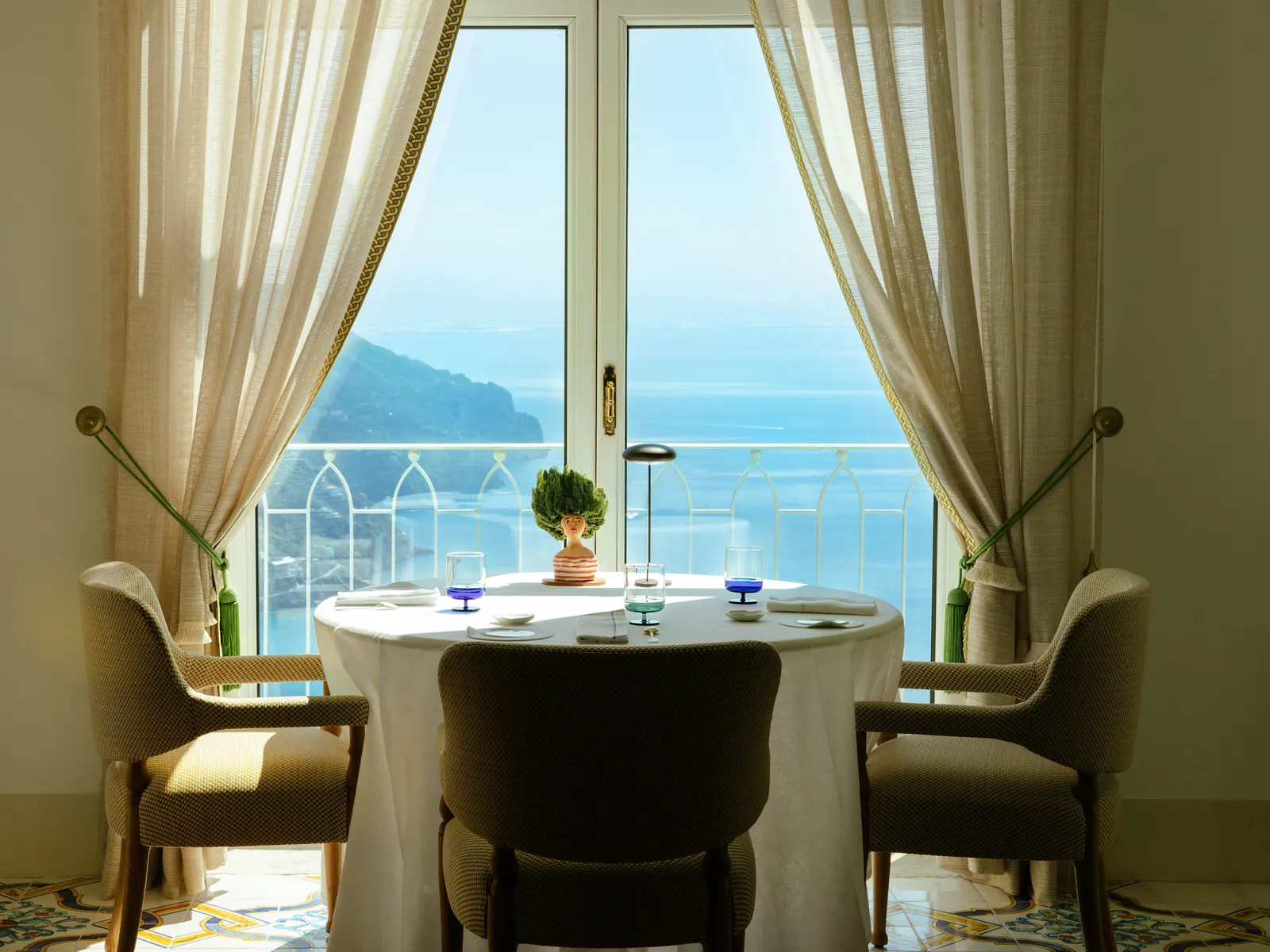 A round laid table before the French window of Il Pantaleone, the curtains tied back and the sea beyond the railing.