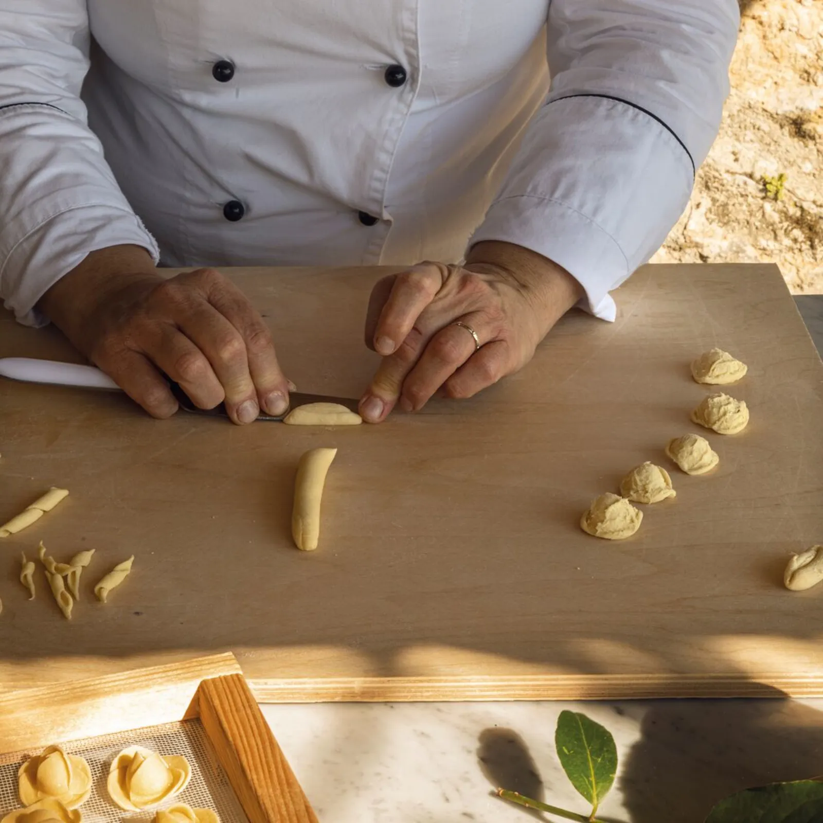 The hands of a cook in a white jacket shaping fresh pasta on a wooden board.
