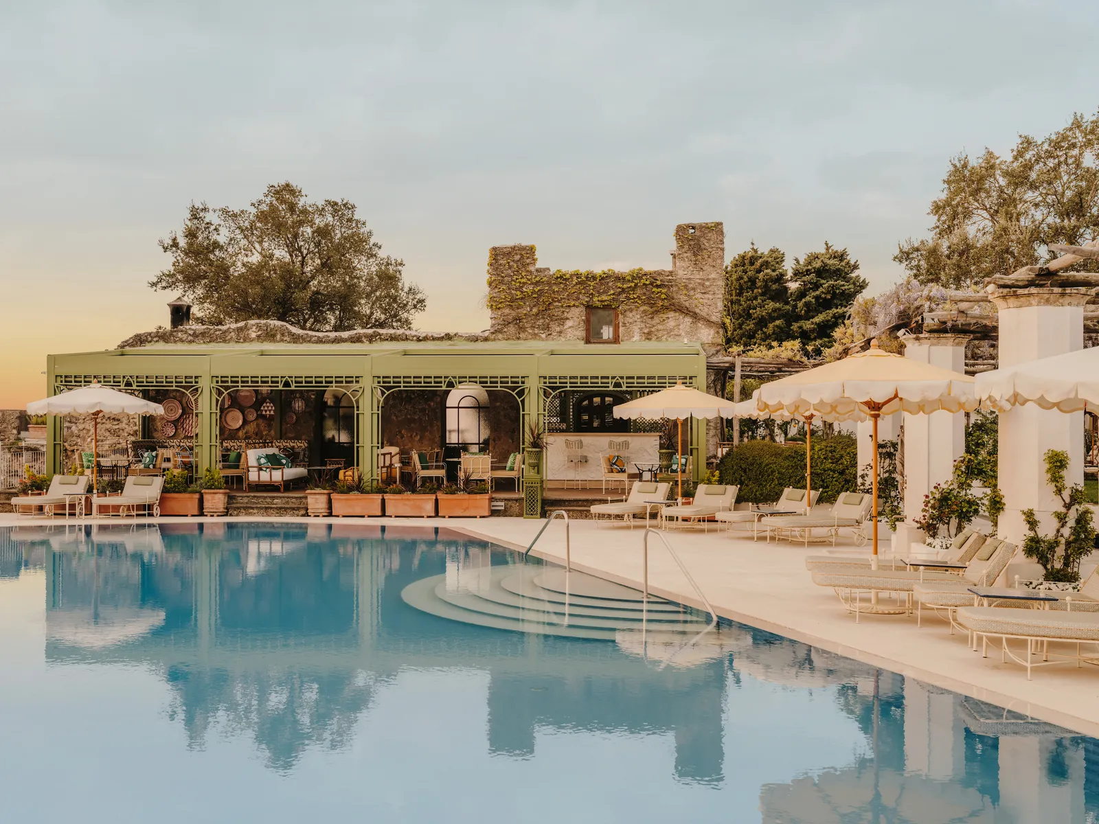 The pool seen from the water: loungers, parasols and the pool club’s green pergola before an old stone wall.