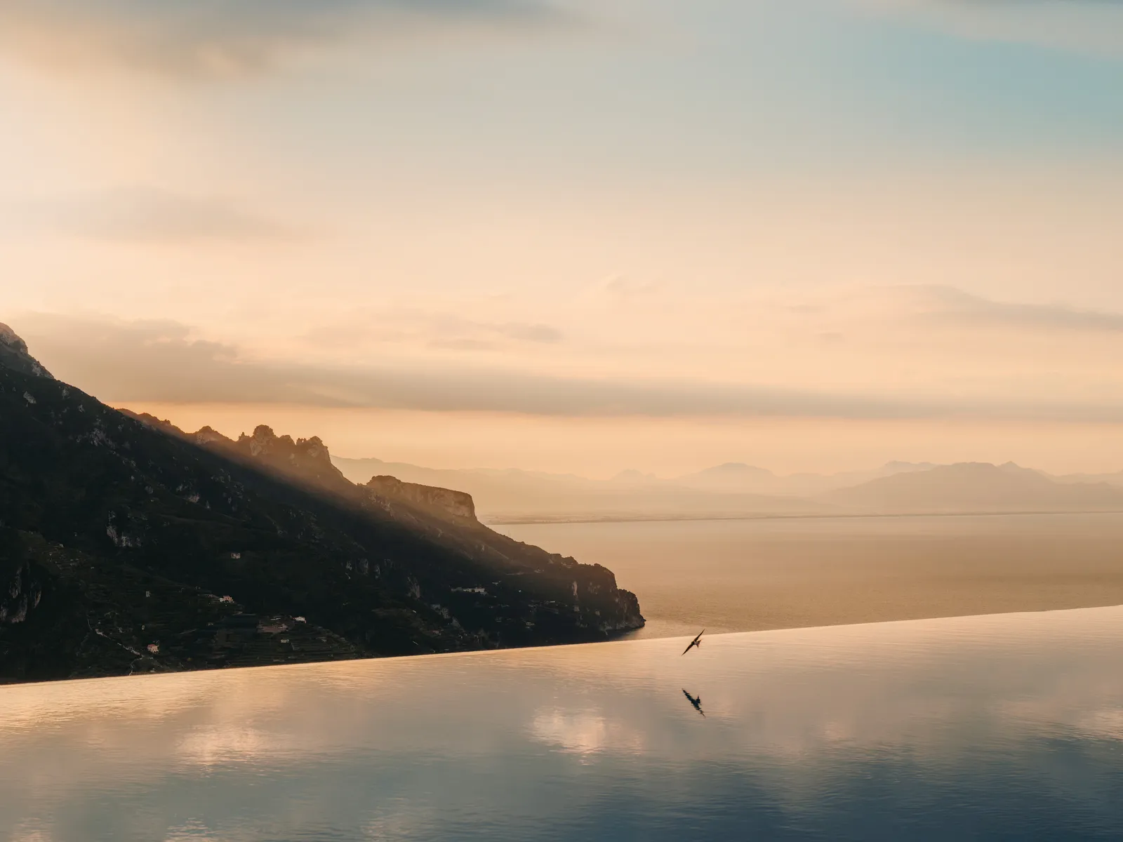 The pool’s infinity edge at sunset, with the dark outline of the coast and a peach-coloured sea.